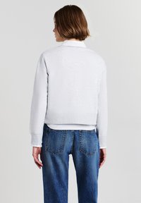 Maglione grigio chiaro, testurizzato, con un design cropped e orlo a costine. Indossato sopra una camicia bianca, abbinato a jeans in denim blu, visto da dietro.