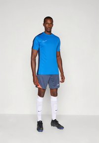 Nike-Sportoutfit mit blauem Kurzarmshirt und grauer Shorts mit orangefarbenem Akzent. Enthält weiße Socken und schwarze Fußballschuhe.
