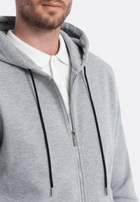 Sweat à zip gris avec une texture douce, cordons noirs et fermeture éclair devant visible. Porté par-dessus une chemise blanche à col.
