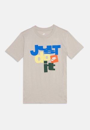 Tricou bej Nike cu mânecă scurtă, având text îndrăzneț și colorat „Just do it” și un mic logo portocaliu Nike în față.