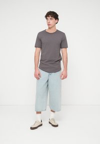 Γκρι κοντομάνικο μπλουζάκι, ανοιχτό μπλε τζιν παντελόνι με φαρδιά μπατζάκια, μπεζ αθλητικά παπούτσια με σκούρες σόλες, στέκεται μπροστά σε απλό λευκό φόντο.