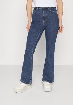 Woman wearing high-waist blue denim jeans with a slight flare and white platform slip-on shoes, standing against a plain background.