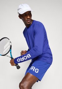 Mėlynas sportinis ilgomis rankovėmis marškinėlis ir šortai su baltais užrašais "ORG". Baltas kepurė ir teniso raketė papildo sportišką išvaizdą.