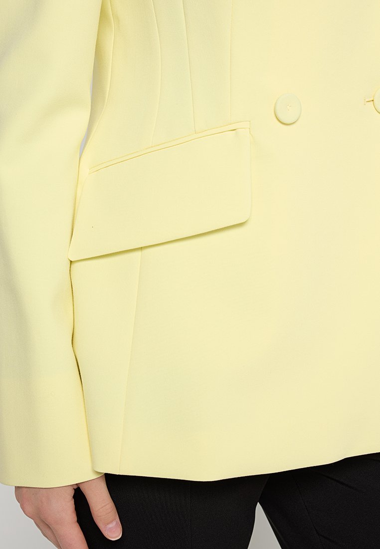 Gelber Blazer aus glattem Stoff mit doppelreihiger Knopfleiste, Reverskragen und einer seitlichen aufgesetzten Tasche.