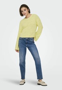 Maglione giallo, strutturato, con scollatura rotonda e maniche a campana, abbinato a jeans dritti blu e mocassini chiari.