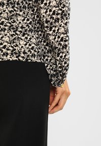 Blusa floreale bianca e nera con maniche lunghe, caratterizzata da un bottone decorativo al polsino e una texture liscia, abbinata a un pantalone nero uniforme.