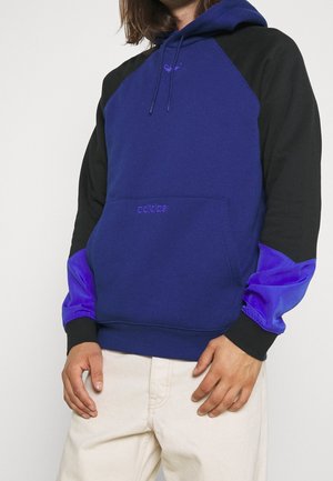 Mand iført en blå og sort Adidas hættetrøje med lilla ærmer og beige bukser, stående mod en ensfarvet hvid baggrund.