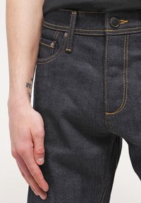 Tmavé džínové kalhoty s žlutým prošitím, vybavené předním kapsou, poutky na opasek a zapínáním na knoflík. Hladká textura, střih na úzkou nohu.