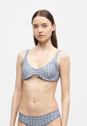 Jeune femme portant un bikini bleu assorti à motifs floraux et géométriques, debout devant un fond clair uni.