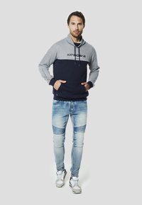 Sudadera con capucha gris y azul marino con bolsillo frontal y capucha con cordón, combinada con vaqueros desgastados de color azul claro y zapatillas multicolores.