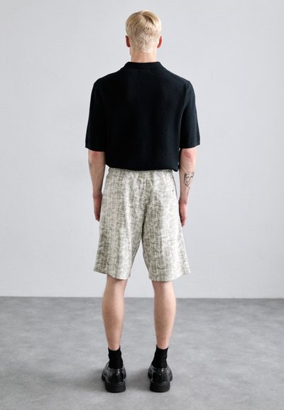 Schwarzes kurzes Strickpolo, kombiniert mit beigen gemusterten Shorts. Das Modell trägt schwarze Socken und glänzende schwarze Schuhe.