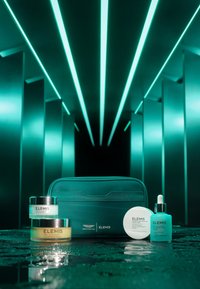 Grønn kosmetisk veske med logo, to kremkrukker, en ansiktsmaske og et serum. Materialene er blanke; krukkene har sølvdetaljer; teksturene varierer.