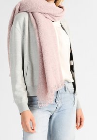 Femme portant un cardigan gris clair zippé, une blouse blanche, un jean bleu clair, et une grande écharpe rose pâle douce enroulée autour du cou.
