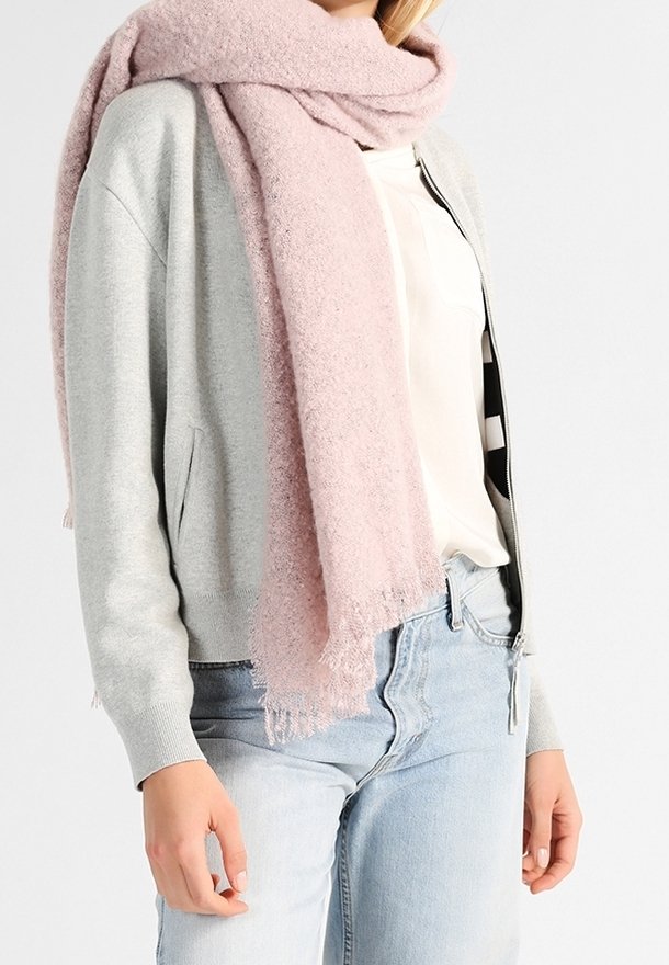 Femme portant un cardigan gris clair zippé, une blouse blanche, un jean bleu clair, et une grande écharpe rose pâle douce enroulée autour du cou.