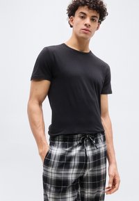 Νεαρός με σγουρά μαλλιά, φορώντας απλό μαύρο μπλουζάκι και μαύρο-λευκό καρό παντελόνι, στέκεται με τα χέρια στις τσέπες.