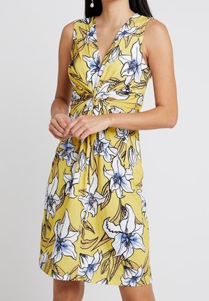 Femme portant une robe jaune sans manches avec un imprimé floral bleu et blanc et un détail de nœud torsadé à la taille.