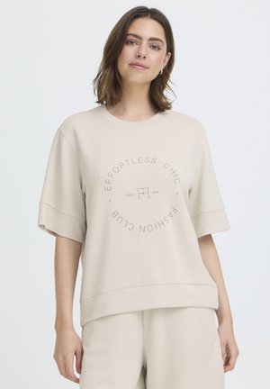 Young woman wearing beige oversized T-shirt with "Effortless Chic Fashion Club" text and matching loose pants against a plain background.