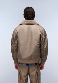 Karmelowa kurtka bomberka z futrzanym kołnierzem, ściągaczami na rękawach i teksturowanym materiałem. Posiada boczne kieszenie i luźny krój.