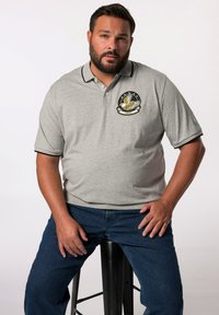 Grå kortärmad polo med svart krage och muddar. Har en märke med en fågel design och text, bärs med blå denimjeans.