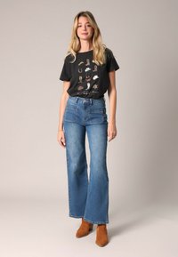 T-shirt noir avec un graphique de chaussures multicolores ; jeans flare taille haute bleus ; bottes à talons chevilles marron clair ; texture lisse.