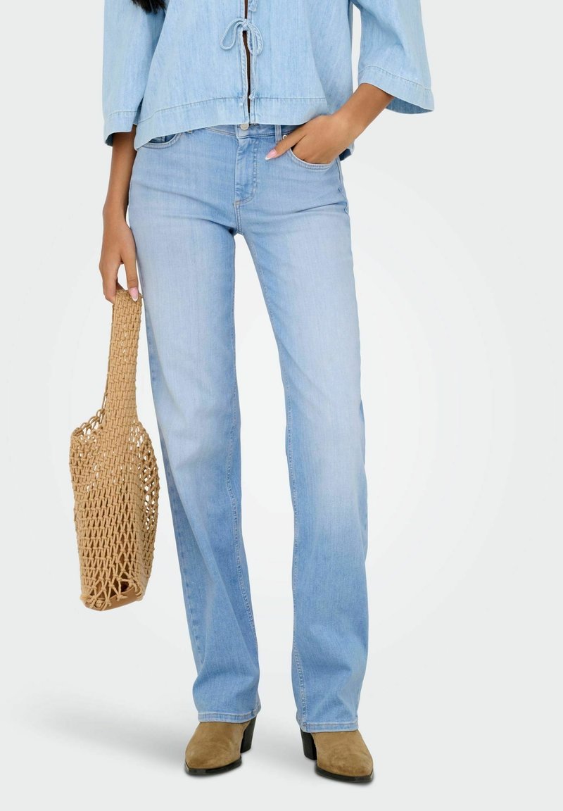 Femme portant un jean bleu clair, une veste en denim claire, des bottes en daim beige et tenant un sac fourre-tout en paille tressée.