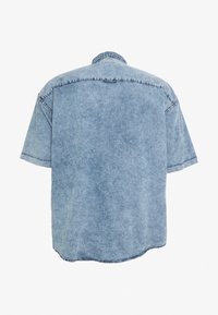 Camisa de denim de manga corta azul clara vista desde atrás, con costuras visibles y un cuello casual, sobre un fondo blanco.