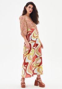 Robe longue avec un haut à manches bouffantes rouge et crème avec un détail en nœud, associée à une jupe colorée à motif ondulé. Talons plateforme marron.