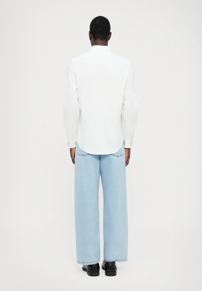 White long-sleeved shirt with a rounded hem, paired with light blue wide-leg jeans and black shoes. Simple design, clean lines, minimal detailing.