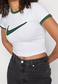 Valkoinen puuvillainen lyhyt paita, jossa on vihreä reunus ja selkeä musta Nike Swoosh -logo. Yhdistetty korkeavyötäröisiin mustiin farkkuihin.