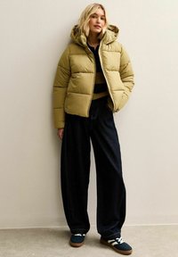 Chaqueta acolchada en verde oliva con capucha forrada, cierre de cremallera y forma cuadrada. Combinada con vaqueros anchos oscuros y zapatillas azules.