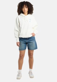 White hoodie with a front pocket and blue logo, paired with denim shorts and white sneakers. Model has curly hair and light socks.