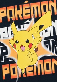 Melna T-krekls ar lielu, dzeltenu Pikachu grafiku un sarkaniem vaigiem. Vārds "Pokémon" ir rakstīts treknos oranžos un baltos burtiem ap dizainu.
