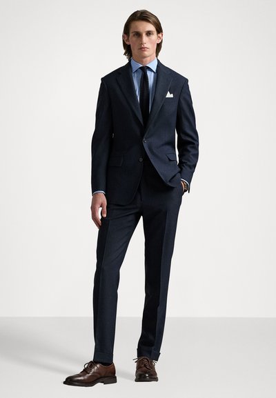 Costume marine ajusté avec des revers nets, deux poches et une texture subtile. Associé à une chemise à carreaux bleu clair et des chaussures habillées marron foncé.