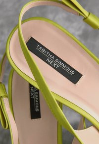 Sandales à talons hauts en satin vert avec une semelle rose, présentant des sangles croisées et une étiquette indiquant "Tabitha Simmons NEXT" en noir.