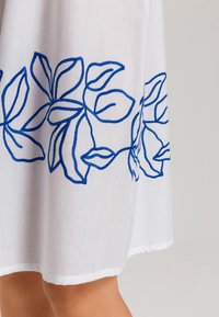 Vestido de tela blanca con hojas bordadas en azul a lo largo del dobladillo. La textura es suave, con un corte recto. Sin adornos adicionales.
