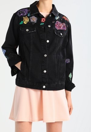 Czarna kurtka dżinsowa z kolorową kwiatową haftem, noszona na jasnoróżowej plisowanej spódnicy, jedna ręka w kieszeni, widok z przodu.