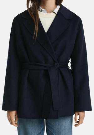 Femme portant un manteau en laine bleu marine ceinturé par-dessus un pull blanc et une chemise à col, associé à un jean bleu clair.
