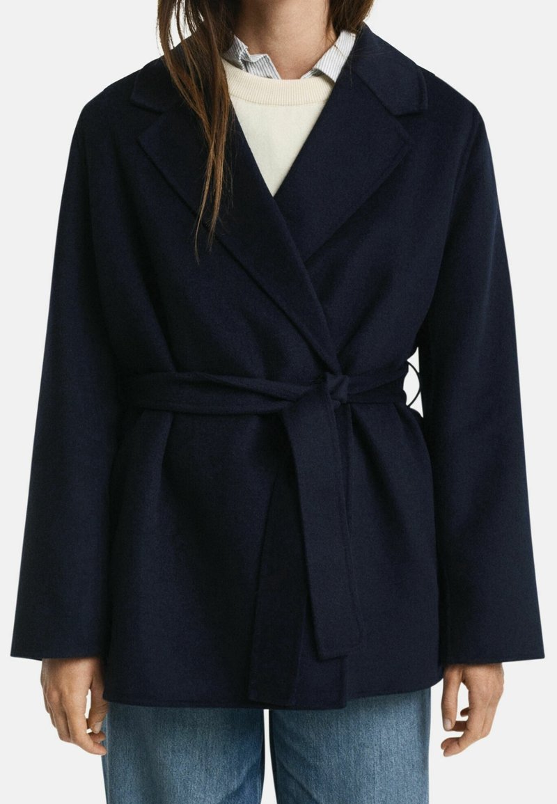 Femme portant un manteau en laine bleu marine ceinturé par-dessus un pull blanc et une chemise à col, associé à un jean bleu clair.