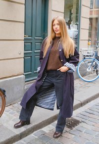 Jeune femme aux longs cheveux blonds porte un manteau sombre, un pull marron, un jean noir et des bottes marron, debout dans une rue pavée près des vélos.