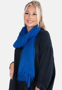 Écharpe bleue texturée en matériau doux, avec des bords effilochés, drapée autour du cou d'une personne portant un haut noir.