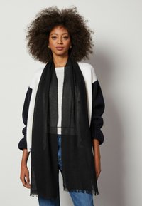 Svart scarf i ett lätt, genomskinligt material med fransade kanter, draperad över en vit topp och en marinblå cardigan, accessoriserad med hoopörhängen.