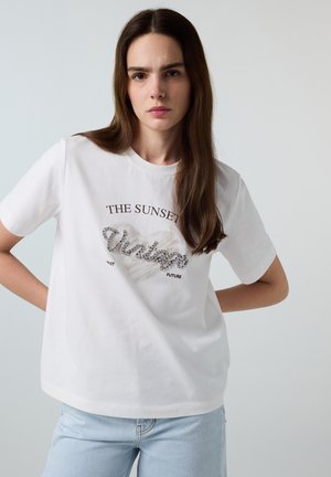 Junge Frau mit langen braunen Haaren, die ein weißes T-Shirt mit dem Schriftzug „THE SUNSET Vintage“ und hellblaue Jeans trägt, steht mit den Händen in der Hüfte.