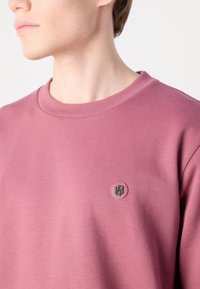 Gros plan d'une personne portant un sweat-shirt mauve à col rond avec un petit patch logo rond sur la poitrine.