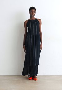 Vestido sem alças, de comprimento total em azul marinho, com riscas verticais; tecido leve e plissado; usado com sapatos planos vermelhos brilhantes.