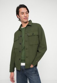 Mees, seljas kihiline roheline särk ja jope, sinistes teksades, seisab ühetaolise valge taustaga, vaadates vasakule.