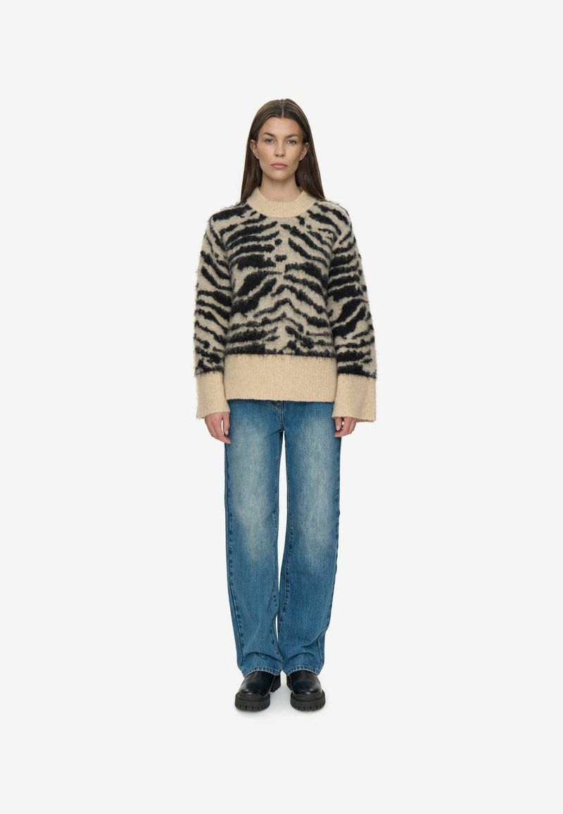 Sort og beige fuzzy sweater med tigerstriber, rund hals og brede manchetter, parret med blå løstsiddende jeans og sort fodtøj.