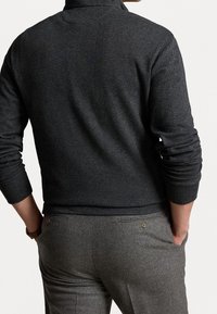 Grauer Fischgrät-Pullover mit hohem Kragen und gerippten Bündchen, kombiniert mit maßgeschneiderten grauen Hosen, die über einen Knopfverschluss an der Gesäßtasche verfügen.