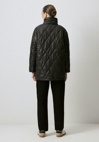 Chaqueta negra acolchada con cuello alto y patrón de diamantes. Combinada con pantalones negros de pierna ancha y zapatos marrones, vista desde la parte de atrás.