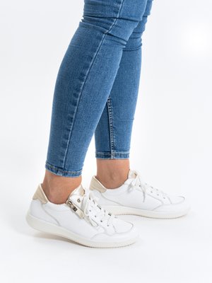 Beine in enganliegenden blauen Jeans und weißen Ledersneakers mit goldenen Akzenten, seitlichem Reißverschluss und weißen Schnürsenkeln auf weißem Hintergrund.