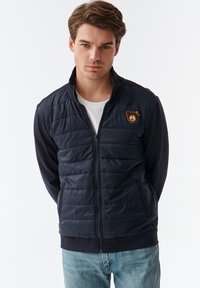 Mørkeblå zip-up jakke med kviltet front, ensfarvede ærmer og et bjørne patch på brystet. Har sidelommer og ribbede manchetter og kant.